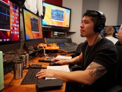 An Asian man sits in front of two computer screens at a desk, wearing a blank t-shirt and black headphones. On the screen he has two black windows open.